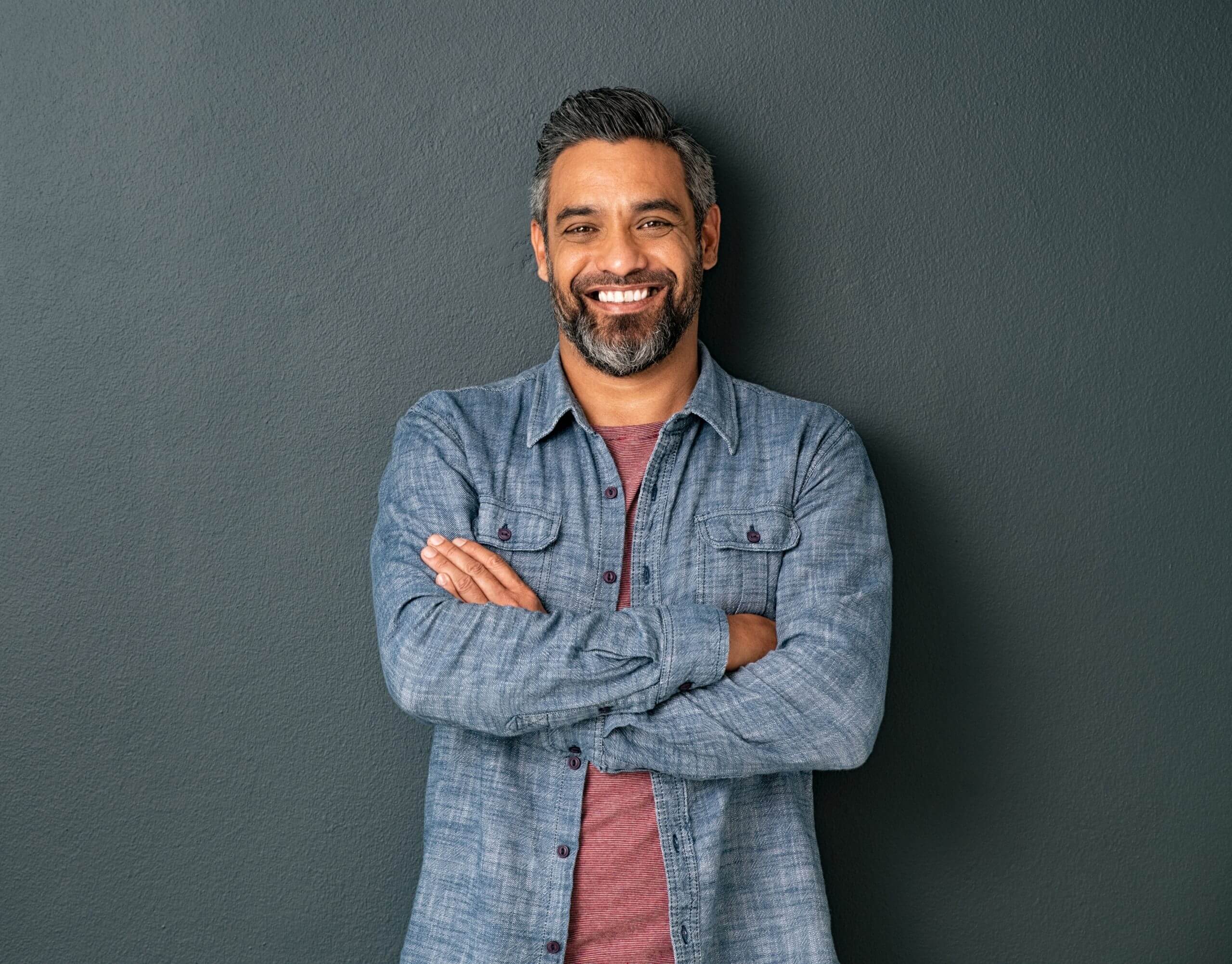 Portrait of smiling middle aged man leaning against a dark grey wall with his arms crossed.