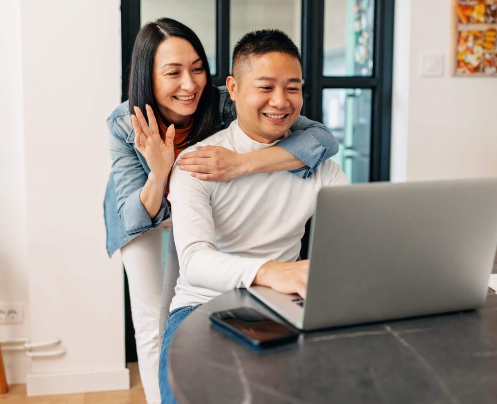 Photo of a man and woman video chatting on a laptop. The man is sitting in a chair and the woman stands behind him with her arm resting around his shoulders.